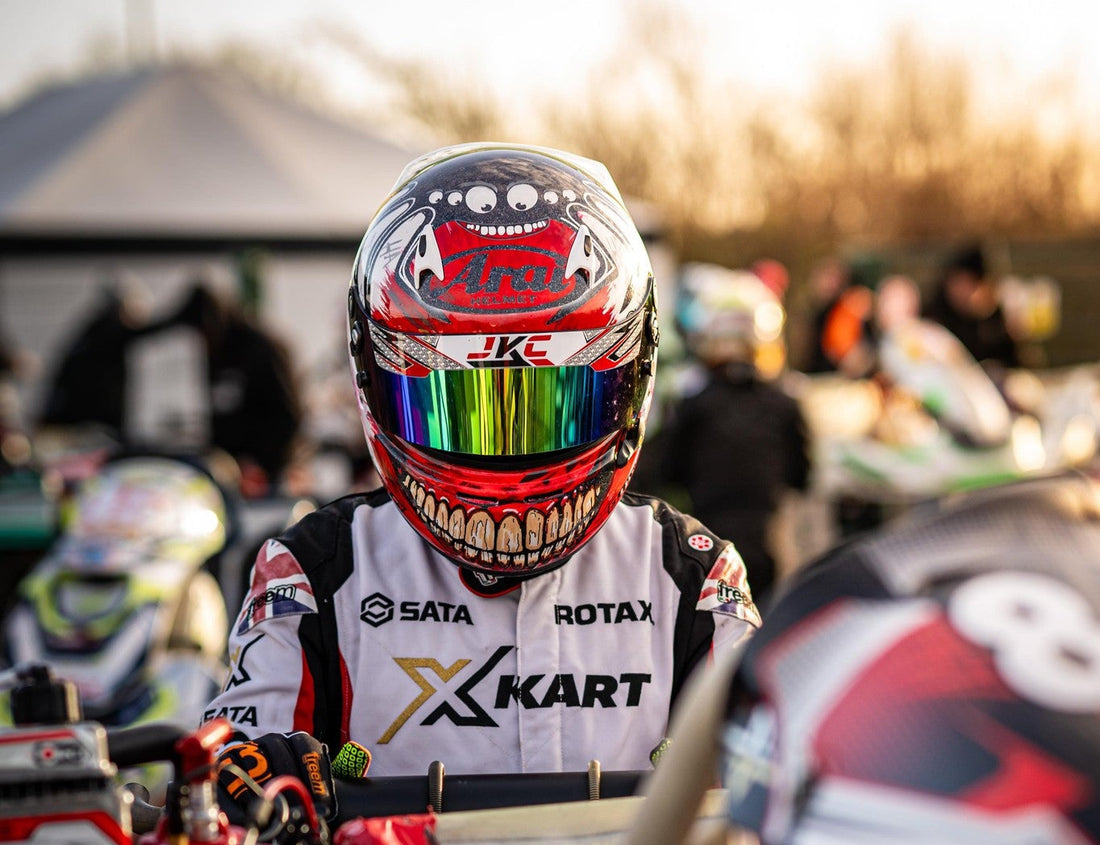 Go-kart driver wearing a custom painted Arai race helmet with a bold teeth design and reflective visor, sitting in kart at the starting grid during a motorsport event.